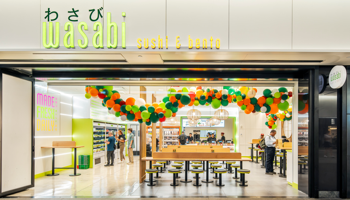 A modern Wasabi sushi and bento restaurant decorated with green, orange, and white balloons. The entrance is open, showing tables, stools, and customers inside. Bright lighting and colorful signage are visible.