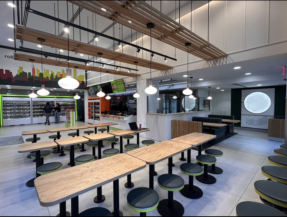 A modern, brightly-lit restaurant interior with wooden tables, round green-accented stools, hanging globe lights, and minimalist decor. A person stands near a wall of refrigerated food displays in the background.